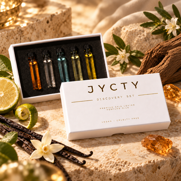 JYCTY Discovery Set premium skin-loving perfume oil, featuring a white box with gold lettering and four sample glass vials inside, styled on a stone surface with limes, vanilla beans, flowers, and amber crystals.