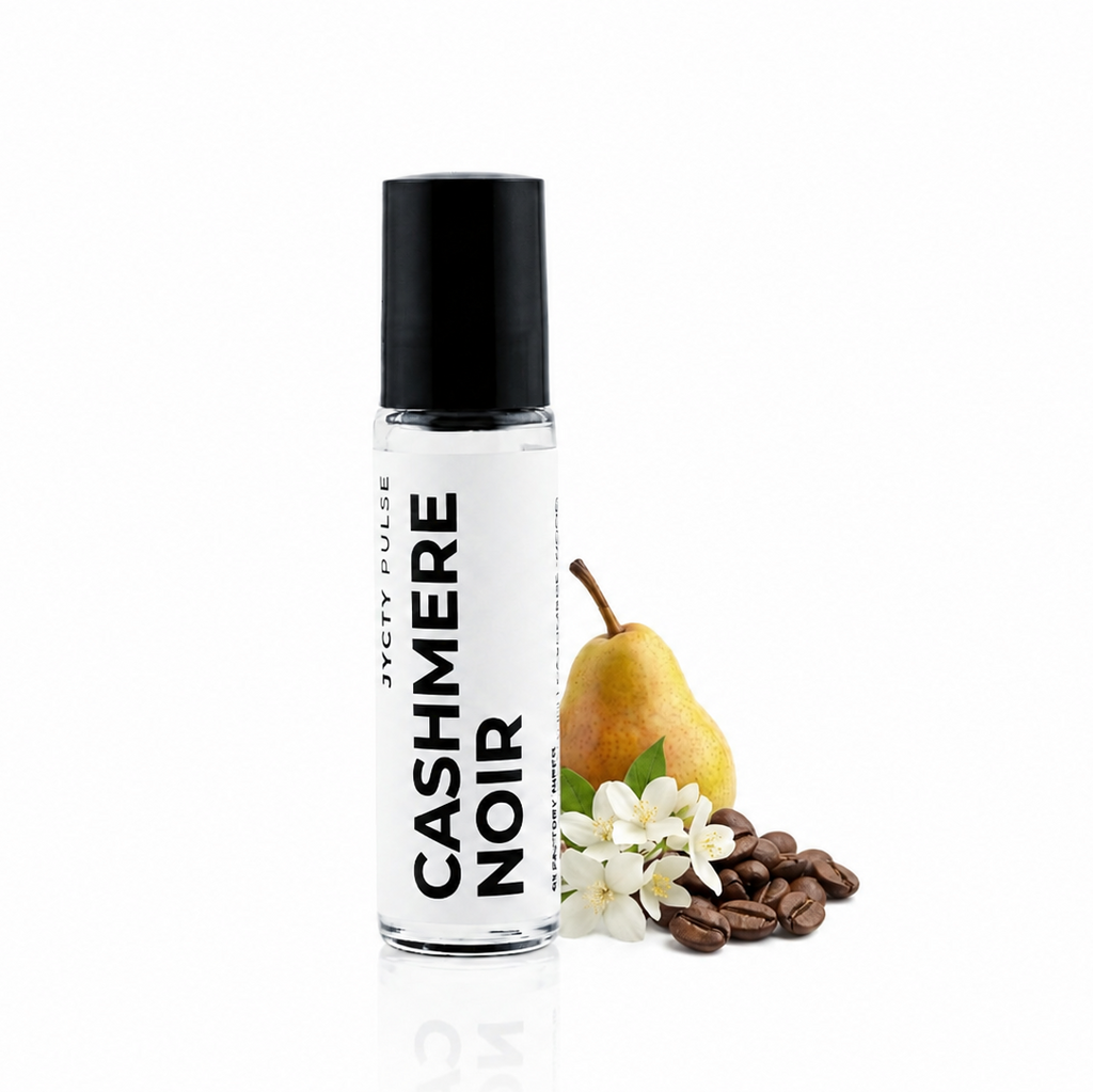 A clear cylindrical bottle with a black cap labeled 'CASHMERE NOIR' stands on a white reflective surface beside a pear, white flowers, and brown coffee beans. The product is shown alone with styled scent-related props on a clean white background.