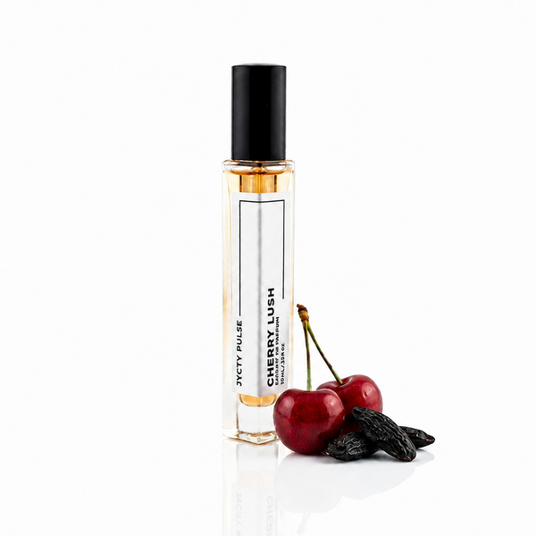 A clear glass perfume roller bottle with a black cap, labeled 'CHERRY LUSH', standing on a white reflective surface next to two red cherries and a dark dried fruit.