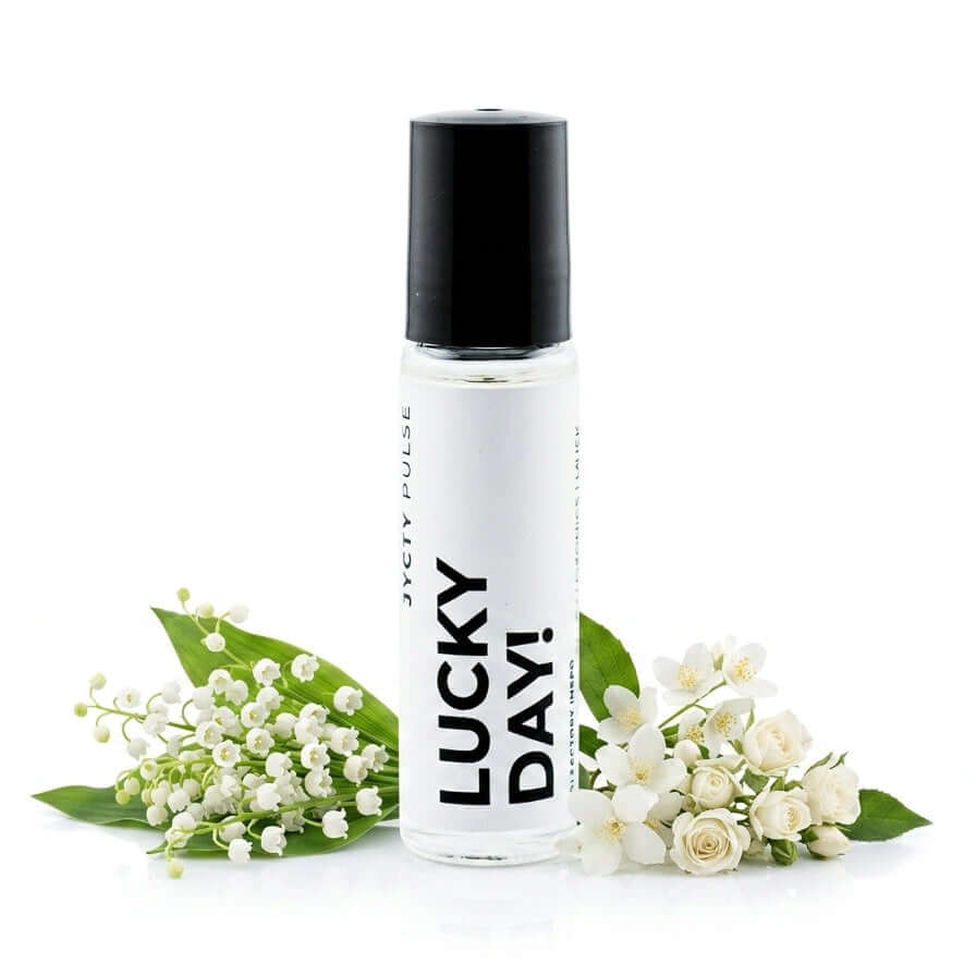 A clear cylindrical roller bottle with a black cap and a white label reading 'LUCKY DAY!' BY JYCTY styled with white flowers and green leaves on a white background.