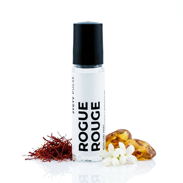 A clear cylindrical roller bottle with a black cap and a white label reading 'ROGUE ROUGE' in bold black text. The bottle is styled on a white reflective surface with red saffron threads, white flowers, and amber gummies around it.