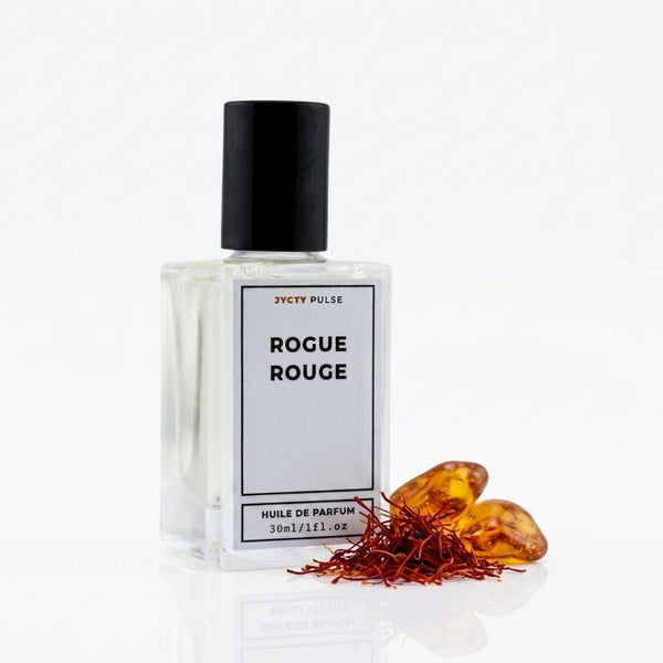 A 30ml bottle of JYCTY PULSE Rogue Rouge PERFUME OIL with a black cap and white label, displayed next to saffron threads and amber saffron flowers on a reflective white surface.