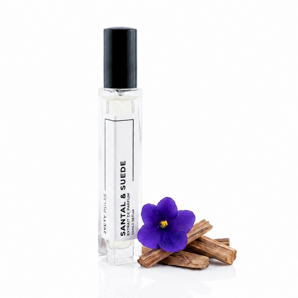 A clear glass perfume roller bottle with a black cap, labeled 'SANTAL & SUEDE' by JYCTY, standing on a white reflective surface next to a purple flower and cinnamon sticks.