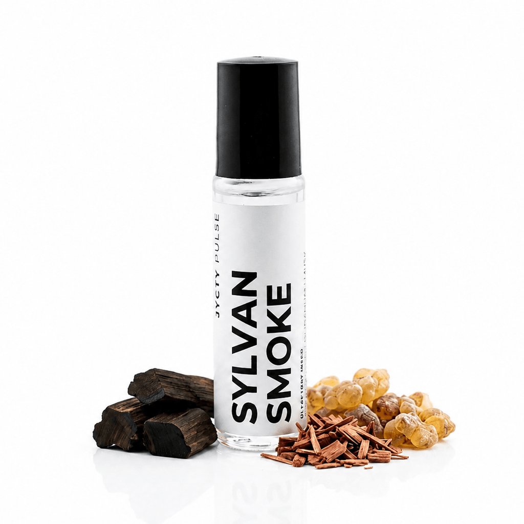 A clear cylindrical bottle with a black cap labeled 'JYCTY PULSE SYLVAN SMOKE' stands on a white background, styled with dark wood pieces, cinnamon sticks, and amber resin pieces at its base.