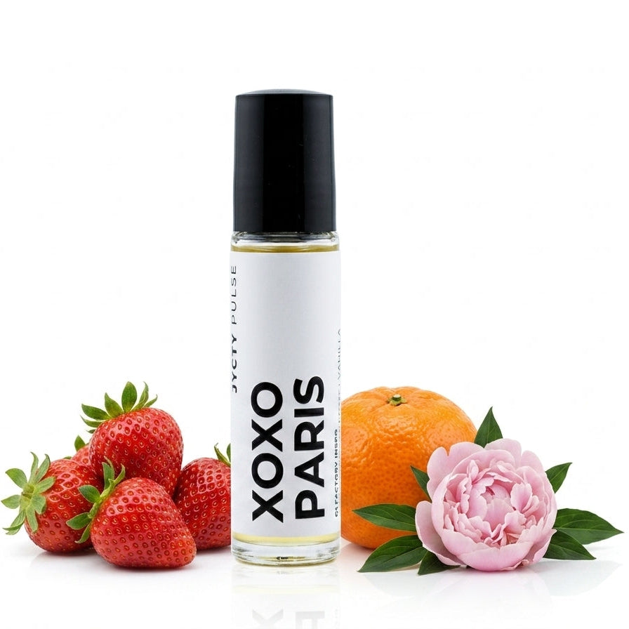 A clear glass roller bottle with a black cap and a white label reading 'XOXO PARIS' stands on a white background, styled with fresh strawberries, an orange, and a pink flower.