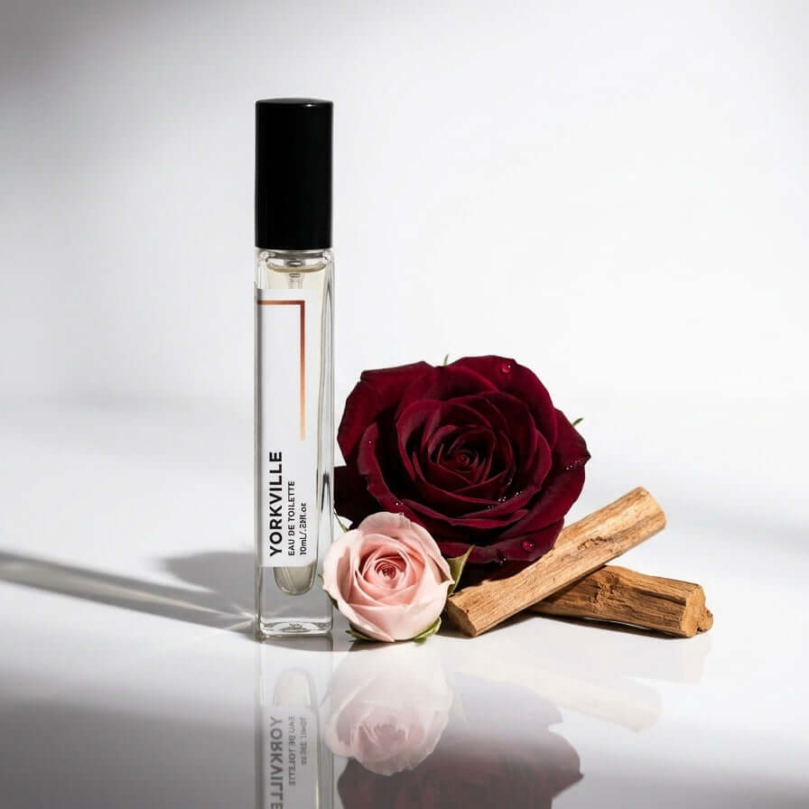 A clear 10ml glass perfume bottle with a black cap stands on a reflective white surface beside two roses, one dark red and one light pink, and two sandalwood. The bottle label reads YORKVILLE.