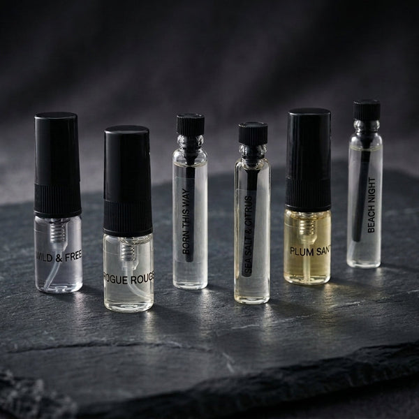Five small perfume sample vials with black caps are arranged on a dark slate surface against a dark studio background. The vials contain clear, pale yellow, and light gold liquids, with labels including Wild & Free, Rogue Rouge, sea saly & citrus, Plum Santal, and Beach Night.